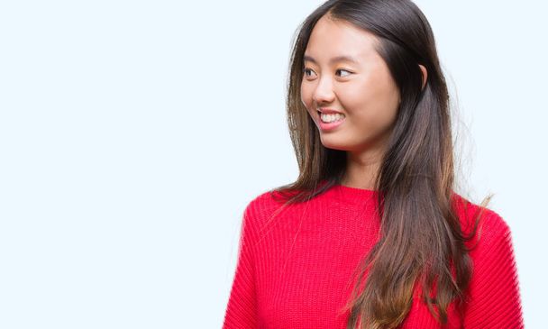 Jonge Aziatische vrouw winter trui dragen over geïsoleerd achtergrond op zoek weg aan kant met glimlach op het gezicht, natuurlijke wijze van uitdrukking. Lachen vertrouwen. - Foto, afbeelding