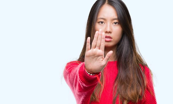 Молодая азиатка в зимнем свитере на изолированном фоне прекращает петь ладонью. Предупреждающее выражение с негативным и серьезным жестом на лице
. - Фото, изображение