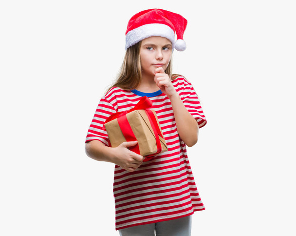 Mooi meisje dragen kerstmuts en bedrijf gift over geïsoleerde achtergrond ernstige gezicht na te denken over de vraag, zeer verward idee - Foto, afbeelding