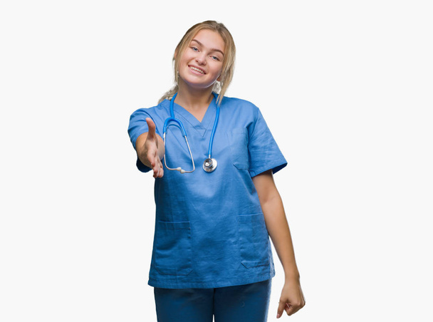 Νεαρός γιατρός καυκάσιος γυναίκα που φοράει στολή χειρουργός πάνω από απομονωμένες φόντο χαμογελώντας προσφέροντας φιλική χειραψία ως χαιρετισμό και καλωσόρισμα. Επιτυχημένη επιχείρηση. - Φωτογραφία, εικόνα