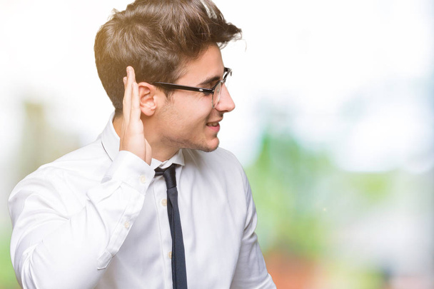 Mladí obchodní muž nosí brýle izolované pozadí s úsměvem s rukou přes ucho naslouchá slyšení zvěsti nebo drby. Hluchota koncept. - Fotografie, Obrázek