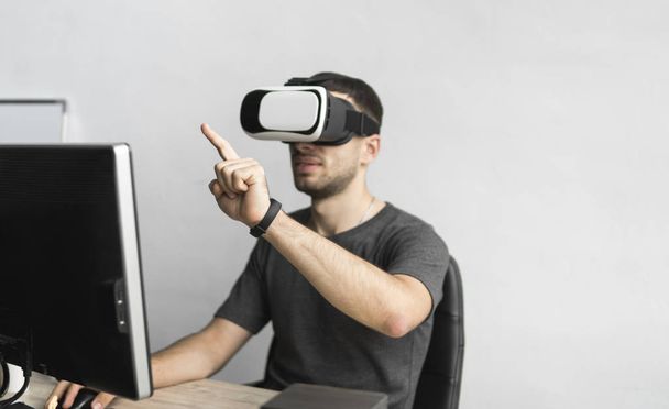 Young man wearing virtual reality goggles headset and sitting in the office against computer. Connection, technology, new generation. Man trying to touch objects or control VR with a hand. - Фото, изображение