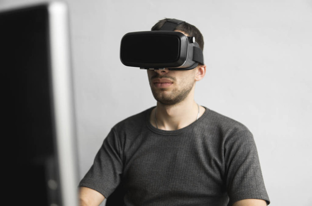 Young man wearing virtual reality goggles headset, vr box and sitting in the office against computer monitor. Connection, technology, new generation, progress concept. - Фото, изображение