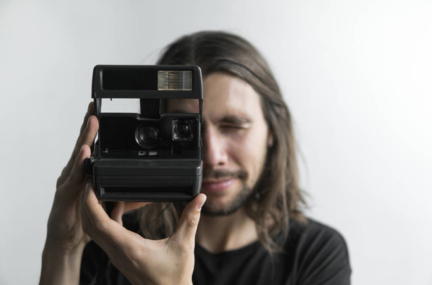 Handsome young bearded man with a long hair and in a black shirt holding vintage old-fashioned film camera on a white background and looking in camera viewfinder. - Foto, immagini