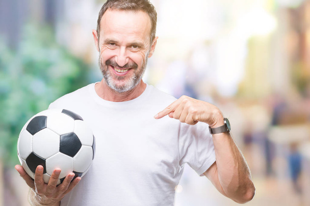 Μέση ηλικία γηραιός ανώτερος άνθρωπος, κρατώντας την μπάλα ποδοσφαίρου ποδοσφαίρου πέρα από το απομονωμένο υπόβαθρο με τεντωμένο έκπληξη πρόσωπο για τον εαυτό του - Φωτογραφία, εικόνα