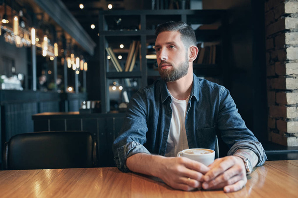 Уверенный в себе человек, наслаждающийся чашкой кофе во время обеденного перерыва в помещении кафе выглядит задумчиво
 - Фото, изображение
