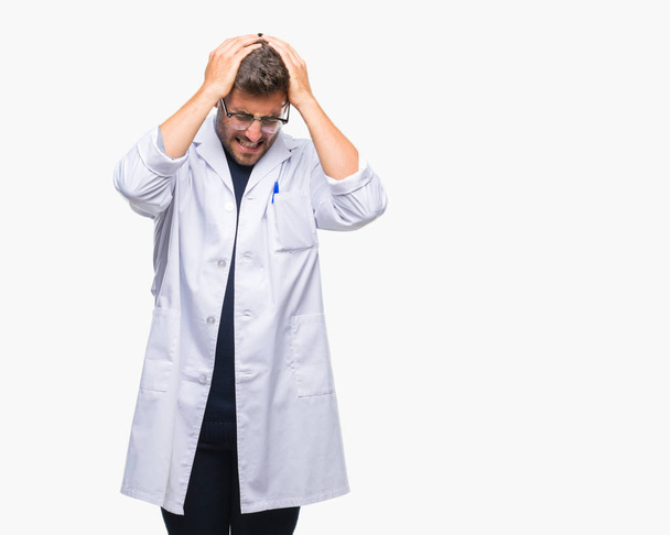Jonge knappe man dragen arts, scientis vacht over geïsoleerde achtergrond lijden hoofdpijn wanhopig en benadrukt omdat pijn en migraine. Handen op het hoofd. - Foto, afbeelding