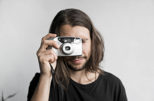 Handsome young bearded man with a long hair and in a black shirt holding vintage old-fashioned film camera on a white background and looking in camera viewfinder. - Zdjęcie, obraz
