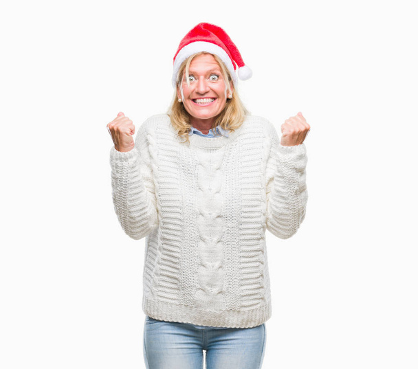 Middle age blonde woman wearing christmas hat over isolated background celebrating surprised and amazed for success with arms raised and open eyes. Winner concept. - Photo, Image