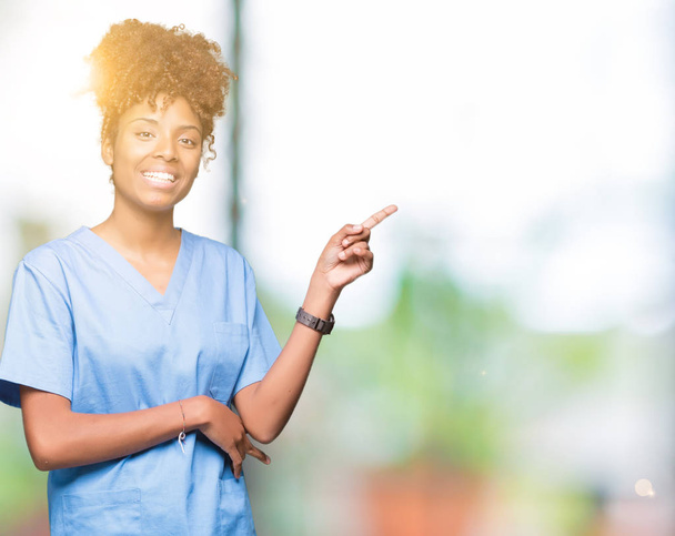 Jeune femme médecin afro-américaine sur fond isolé avec un grand sourire sur le visage, pointant avec la main et le doigt vers le côté en regardant la caméra
. - Photo, image