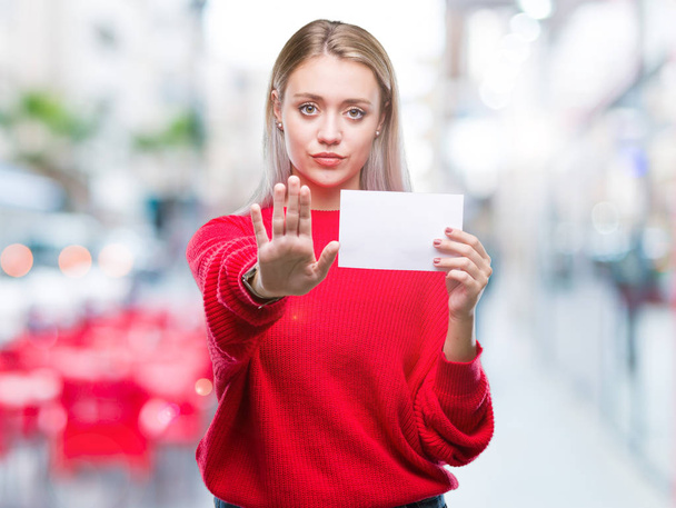 Νεαρή ξανθιά γυναίκα κρατώντας κάρτα κενό χαρτί πάνω από το απομονωμένο φόντο με άνοιγμα πλευρά κάνει στοπ με σοβαρές και αυτοπεποίθηση έκφραση, χειρονομία άμυνα - Φωτογραφία, εικόνα