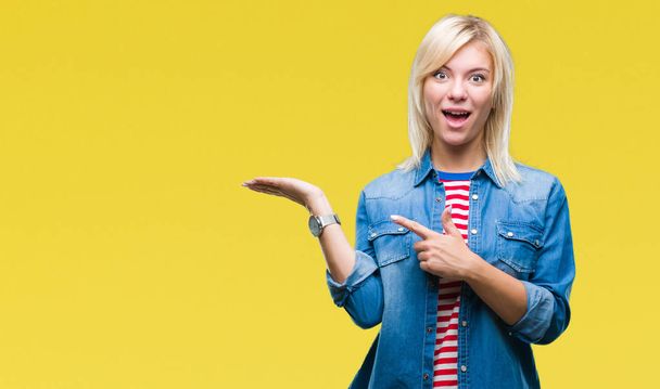 Young beautiful blonde woman wearing denim jacket over isolated background amazed and smiling to the camera while presenting with hand and pointing with finger. - Photo, Image