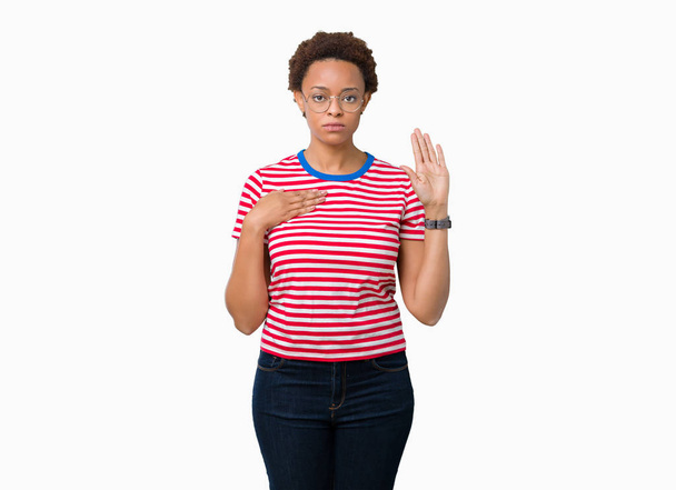 Piękna młoda kobieta Afroamerykanów w okularach na na białym tle przeklinanie ręką na klatce piersiowej i otwartej dłoni, co przysięgą obietnicy lojalności - Zdjęcie, obraz