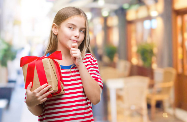 Young beautiful girl giving christmas or valentine gift over isolated background serious face thinking about question, very confused idea - Fotó, kép