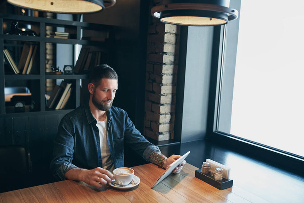Young man drinking coffee in cafe and using tablet computer - 写真・画像