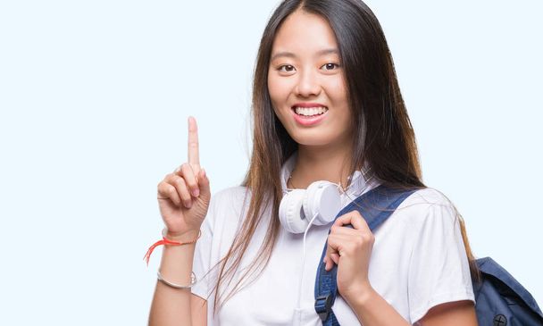 Young asian woman wearing backpack and headphones over isolated background surprised with an idea or question pointing finger with happy face, number one - Foto, Bild
