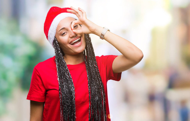 Joven chica afroamericana trenzada con sombrero de Navidad sobre fondo aislado haciendo un gesto bien con la mano sonriente, los ojos mirando a través de los dedos con la cara feliz
. - Foto, Imagen
