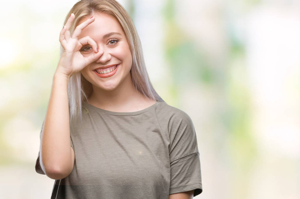 Young blonde woman over isolated background doing ok gesture with hand smiling, eye looking through fingers with happy face. - Φωτογραφία, εικόνα