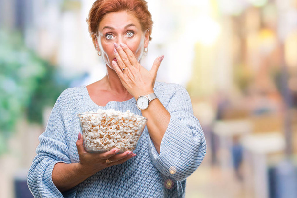 Atrractive senior caucasian redhead woman eating popcorn over isolated background cover mouth with hand shocked with shame for mistake, expression of fear, scared in silence, secret concept - Photo, Image