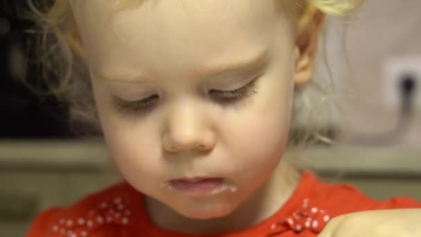 Μικρό χαριτωμένο μωρό κορίτσι με σγουρά μαλλιά και γκρίζα μάτια, ντυμένη με ένα κόκκινο φόρεμα με λευκό πουά, τρώει κρέμα μους - Πλάνα, βίντεο