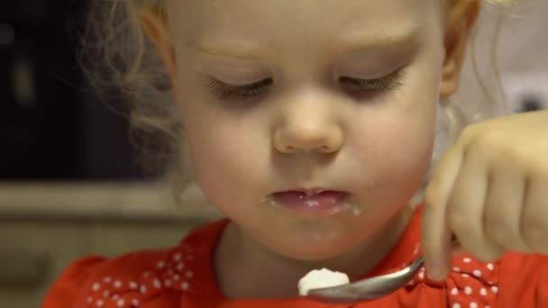 Little cute baby girl with curly hair and gray eyes, dressed in a red dress with white polka dots, is gently tasting cream mousse - Footage, Video