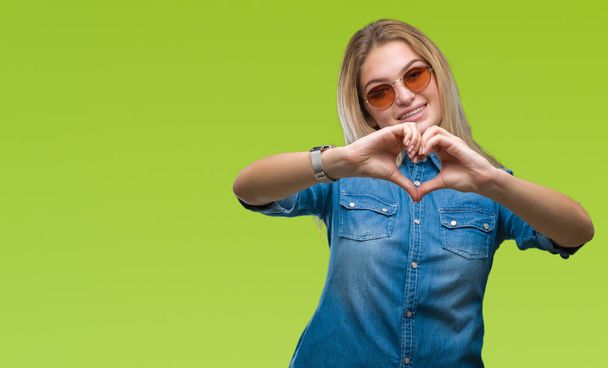 Jonge Kaukasische vrouw dragen van een zonnebril op geïsoleerde achtergrond glimlachend in liefde met hartsymbool en vorm met de handen. Romantische concept. - Foto, afbeelding
