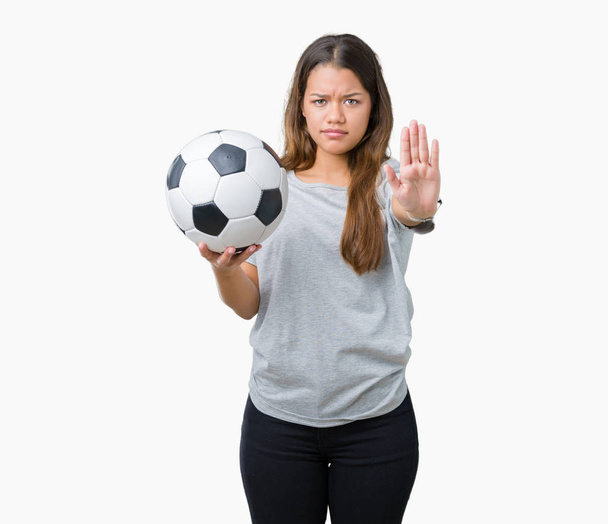 Молодая красивая брюнетка женщина держит футбольный мяч на изолированном фоне с открытой рукой делает знак стоп с серьезным и уверенным выражением, защитный жест
 - Фото, изображение