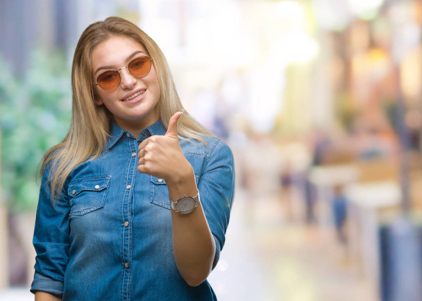 Νεαρή γυναίκα Καυκάσιος φορώντας γυαλιά ηλίου πάνω από το απομονωμένο φόντο κάνει ευτυχισμένη μπράβο χειρονομία με το χέρι. Την έγκριση έκφραση κοιτάζοντας την κάμερα με δείχνει επιτυχία. - Φωτογραφία, εικόνα