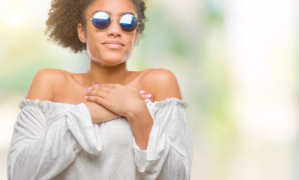 Nuori afroamerikkalainen nainen aurinkolasit päässä eristetty tausta hymyilee kädet rinnassa suljetuin silmin ja kiitollinen ele kasvoilla. Terveyskäsite
. - Valokuva, kuva