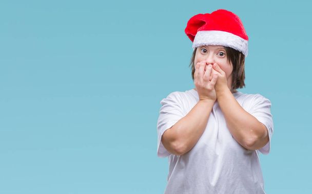 Νεαρή ενήλικη γυναίκα με σύνδρομο down που φοράει καπέλο Χριστουγέννων πέρα από το απομονωμένο υπόβαθρο σοκαρισμένος που καλύπτουν το στόμα με τα χέρια για το λάθος. Μυστική έννοια. - Φωτογραφία, εικόνα
