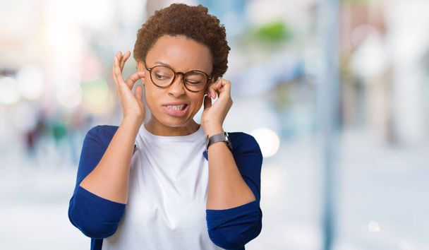 Joven mujer afroamericana hermosa con gafas sobre el fondo aislado cubriendo las orejas con los dedos con expresión molesta por el ruido de la música fuerte. Concepto sordo
. - Foto, imagen
