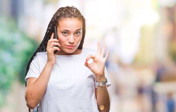 Joven trenzado pelo afroamericano chica mostrando llamando usando teléfono inteligente sobre fondo aislado haciendo signo de ok con los dedos, excelente símbolo
 - Foto, Imagen
