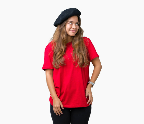 Jonge mooie brunette fashion vrouw dragen rode t-shirt en zwarte baret over geïsoleerde achtergrond op zoek weg aan kant met glimlach op het gezicht, natuurlijke wijze van uitdrukking. Lachen vertrouwen. - Foto, afbeelding