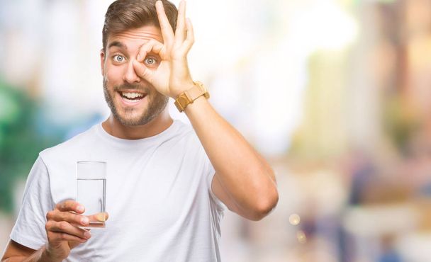 Jonge knappe man glas water drinken op geïsoleerde achtergrond met blij gezicht lachend doen ok teken met de hand op oog kijken door de vingers - Foto, afbeelding