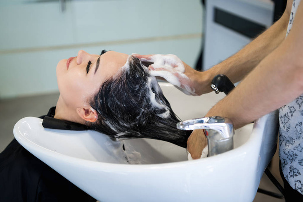 Beauty and people concept - happy young woman with hairdresser washing head at hair salon - Foto, Imagem