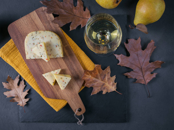A glass of white wine was served with cheese in a cutting board on dark background. Autumn picnic with cheese, wine and dry leaves in rustic style. Overhead - Foto, afbeelding