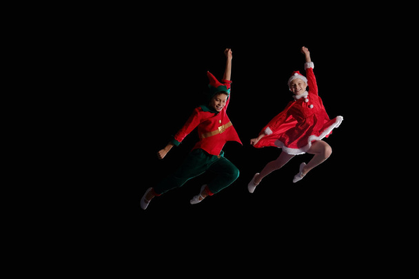 Christmas time, childhood, fairy tale. Cheerful kids - A young girl wearing a Santas costume and boy wearing elf costume flying together - Photo, Image