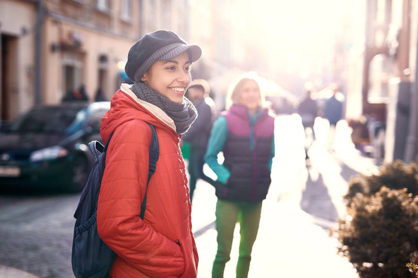 молодая улыбающаяся туристка в красной куртке, сером шарфе и кепке гуляет с друзьями по улице старого Львова в солнечный осенний или весенний день
 - Фото, изображение