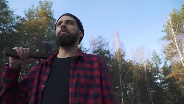 Bearded guy stands with an axe in his hand in the forest.. Brutal serious man with axe outdoors. - Séquence, vidéo