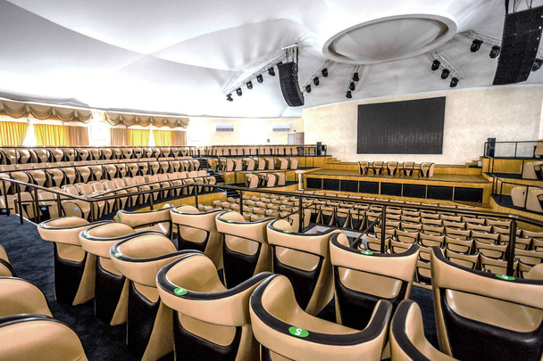 Interior luminoso de una moderna sala de conciertos con un escenario y filas de cómodas sillas de cuero, un escenario y sistema de amplificación acústica bajo luz natural, preparado para la presentación
 - Foto, Imagen