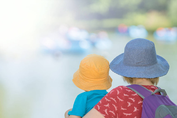 Caucasian woman wearing blue hat holding her little son wearing yellow hat  in a park in summer - Fotografie, Obrázek