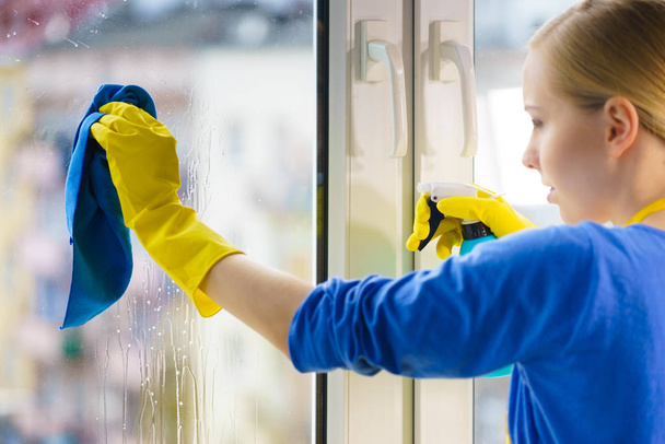 Young woman in yellow gloves cleaning window pane at home with rag and spray detergent. Cleaning concept - 写真・画像