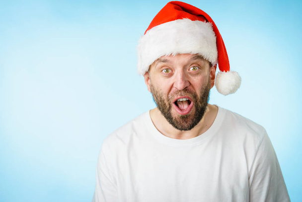 Bearded man wearing santa claus hat surprised expression, side view on blue with copy space. Christmes time. - Photo, Image
