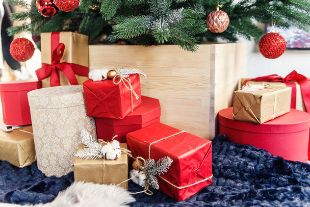 Packed in colored kraft paper holiday gift boxes under the Christmas tree. Red, gold and brown boxes near Christmas tree. - Фото, изображение