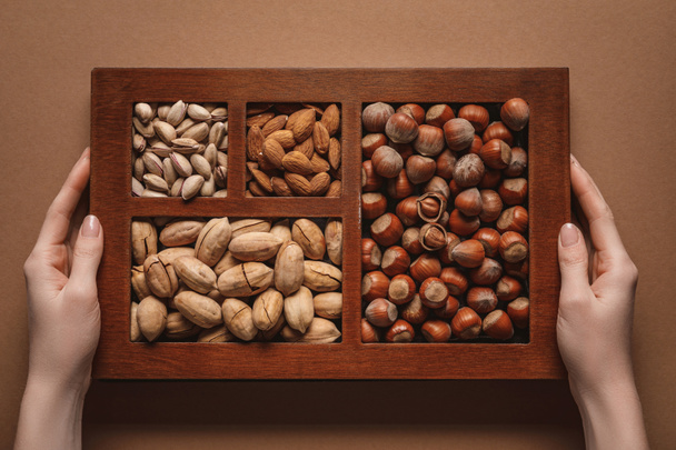 partial view of woman holding box with assortment of different nuts on brown background - Фото, изображение