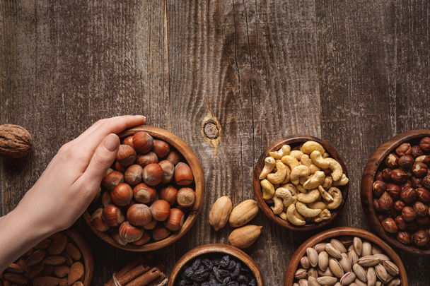 partial view of woman holding bowl with hazelnuts on wooden tabletop with different nuts around - Fotoğraf, Görsel