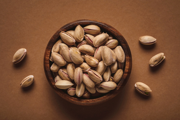 top view of pistachio nuts in wooden bowl on brown backdrop - Fotó, kép