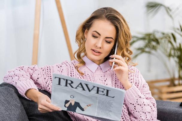 attractive woman sitting on sofa, talking on smartphone and reading travel newspaper in living room - Photo, image