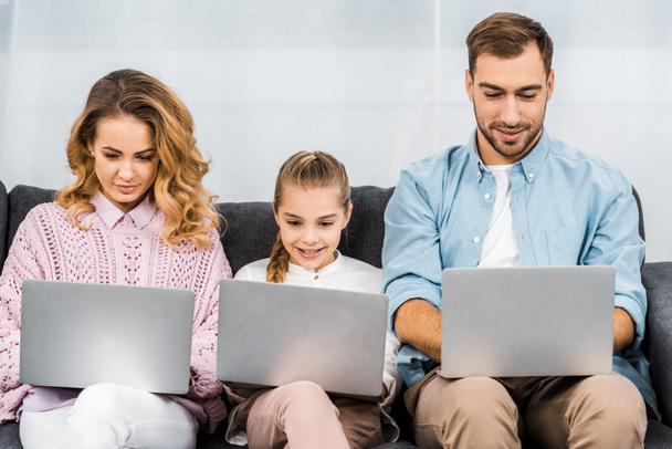 cute girl with two parents using laptops and sitting on sofa in living room - Fotografie, Obrázek