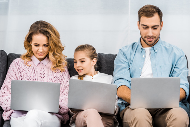 cute girl with two parents sitting on sofa and using laptops in apartment - Photo, Image